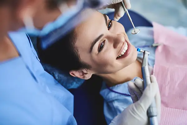 A woman smiling while undergoing a dental cleaning procedure performed by a dentist in a clinic.